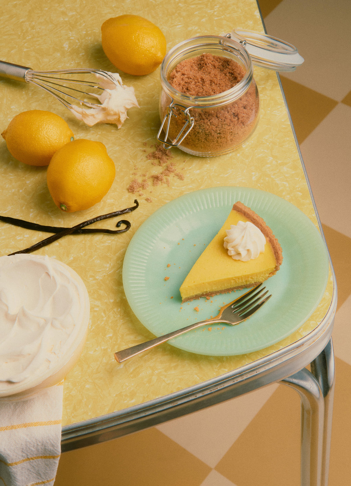 Slice of lemon cake with whipped cream on a green plate, surrounded by lemons, a jar of cinnamon, and vanilla beans on a yellow table.